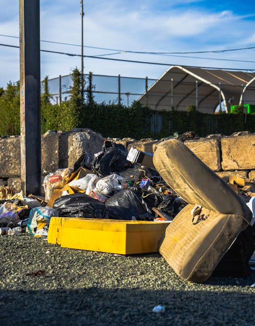 A close-up view of a pile of discarded rubbish on a gravel surface outdoors, consisting mainly of black plastic trash bags filled with waste, some partially torn open revealing packaging and household waste, alongside an old, overgrown, and worn yellow plastic container lying on its side. In the background, a low stone wall separates the rubbish from a hedge and a sports court with a partially visible metal fence and a pergola structure with a curved roof. Overhead, electrical wires stretch across the sky, which is partly cloudy with patches of blue. The scene captures an outdoor area that appears to be part of a private or informal waste disposal site, reflecting the context of rubbish removal services provided by Rubbish Collection Wandsworth, suitable for alternative waste handling or on-site clearance scenarios.