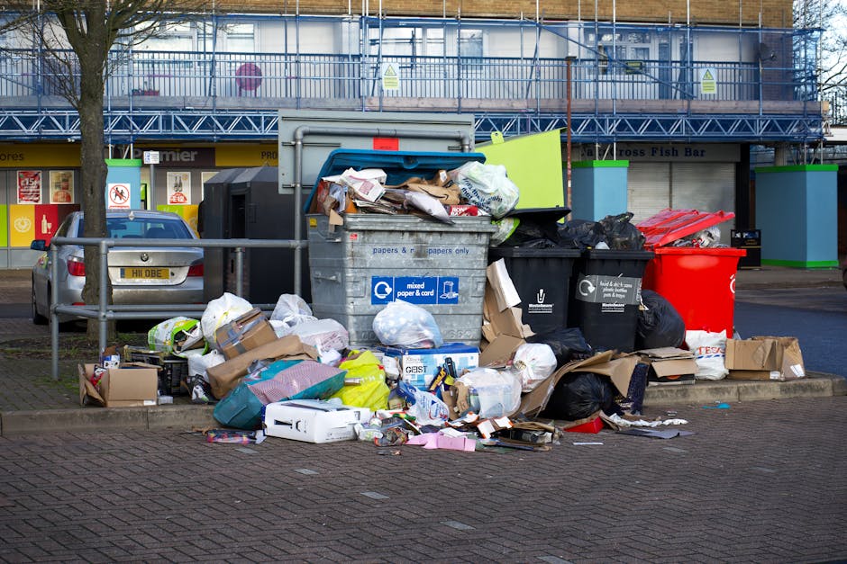A collection of overflowing waste bins and scattered rubbish around a dedicated recycling area situated on a paved urban street. The stop-and-go refuse includes large black garbage bags, flattened cardboard boxes, discarded paper and food packaging, and plastic containers, all spilling onto the ground. A prominent grey mixed-paper-and-cardboard recycling bin, partially open with waste protruding, is surrounded by additional black and red bins meant for general waste and recycling. To the left, a silver hatchback car is parked, with a green metal railing separating the waste area from the rest of the street. The background features a commercial building under renovation, covered in blue scaffolding and protective sheeting, with shopfronts visible underneath. The area appears to be a designated waste disposal point for local businesses or residents, possibly managed by a private waste collection service, illustrating typical urban rubbish accumulation without recent collection. The scene is lit naturally, suggesting daytime, with no signs of immediate waste removal, highlighting the need for waste management services such as those provided by Rubbish Collection Wandsworth.
