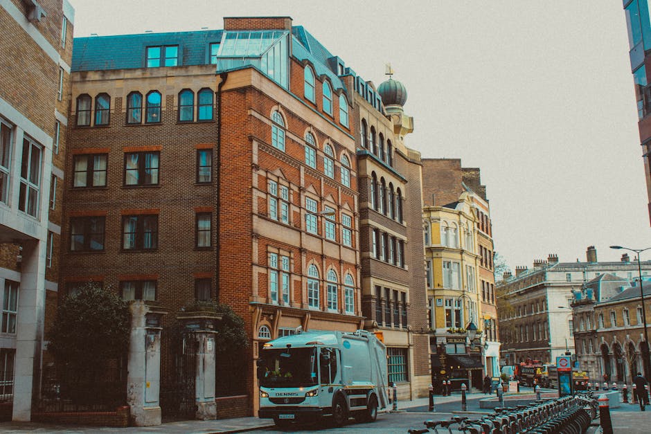 A street scene in an urban area with a focus on a collection truck operated by Rubbish Collection Wandsworth parked along the curb in front of a row of multi-story buildings with Victorian and modern architectural styles. The collection vehicle is light blue with a compact, boxy design and visible side mirrors, positioned on a paved road with bollards and a bike rack in the foreground. Surrounding buildings display various materials, including red brick and light-colored stone, with numerous windows, some with arched or rectangular designs, and decorative façade elements. In the background, other cityscape buildings feature diverse roof types, including a dome-topped tower and a sloped metal roof. The scene is illuminated with daylight, creating a neutral and professional atmosphere typical of a waste collection operation in a busy city environment, illustrating an example of private rubbish removal services on a typical street in central London.