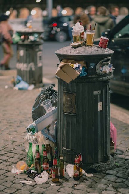 A black, cylindrical public waste bin made of metal with a domed lid, positioned on a cobbled sidewalk. The lid is partially open with various discarded items resting on top, including crumpled paper cups, plastic cups with straws, and a small red container. Several empty glass bottles, mostly brown and green, are scattered on the ground in front of the bin, some upright and others lying on their sides, along with crumpled paper and other debris. A black trash bag sits next to the bin, filled with additional waste. In the background, there is a blurred street scene with pedestrians, moving vehicles, and other upright trash bins, indicating an urban setting. The scene appears to be in the evening or a dimly lit area, with natural and artificial lighting creating a muted atmosphere. The image reflects common issues of littering and the importance of waste management services like those offered by Rubbish Collection Wandsworth.